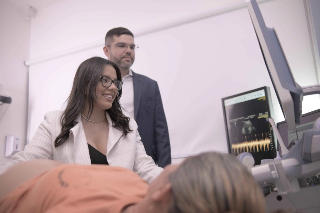 M&eacute;dica realizando exame de ultrassonografia obst&eacute;trica em paciente, com m&eacute;dico observando ao fundo.
