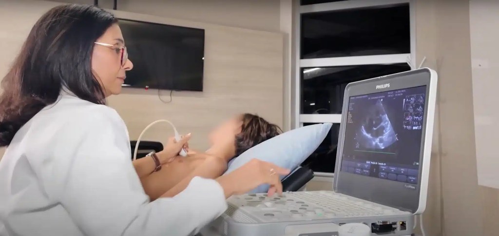 Médica realiza ecocardiograma em uma criança deitada, com a imagem do exame visível na tela do equipamento.