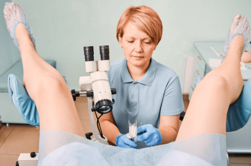 Ginecologista realizando exame ginecol&oacute;gico em uma paciente deitada em posi&ccedil;&atilde;o ginecol&oacute;gica, com as pernas apoiadas em suportes. A m&eacute;dica usa luvas azuis e manipula um esp&eacute;culo vaginal, com colposc&oacute;pio posicionado para visualiza&ccedil;&atilde;o detalhada do colo do &uacute;tero. Ambiente cl&iacute;nico, limpo e iluminado.