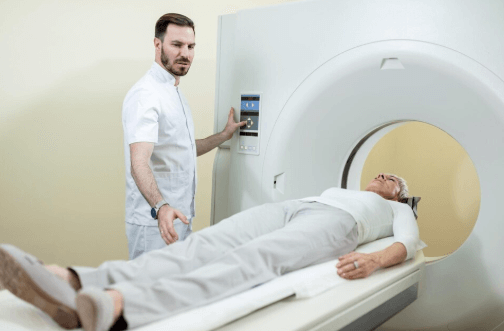 Profissional operando um equipamento de tomografia computadorizada enquanto uma paciente idosa est&aacute; deitada na mesa do aparelho, prestes a realizar uma angiotomografia de coron&aacute;rias.