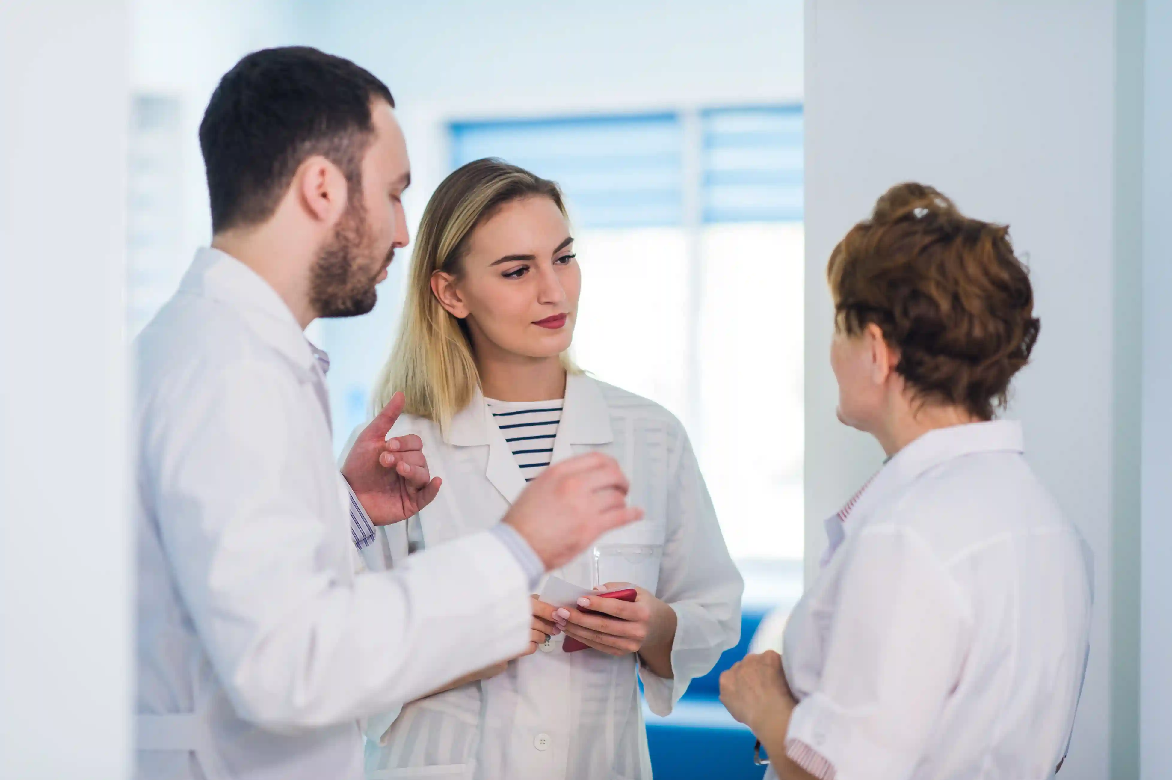 Tr&ecirc;s profissionais de sa&uacute;de em conversa no corredor de um hospital, usando jalecos brancos.
