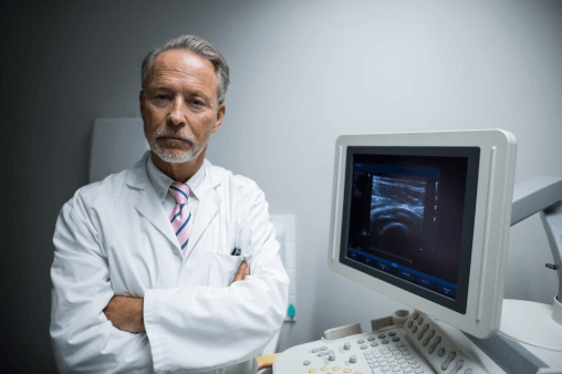 A imagem mostra um m&eacute;dico, de bra&ccedil;os cruzados, ao lado de uma m&aacute;quina de ultrassonografia.