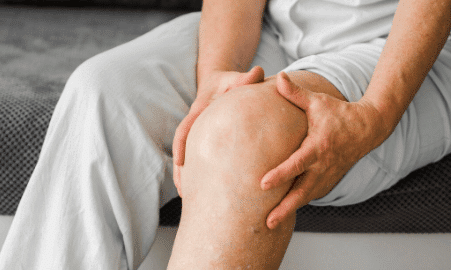 Pessoa sentada segurando o joelho com as duas m&atilde;os, demonstrando dor ou desconforto na articula&ccedil;&atilde;o.
