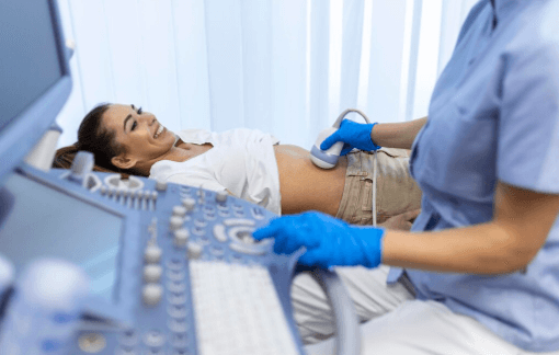 Mulher deitada em maca sorrindo enquanto profissional de sa&uacute;de realiza exame de ultrassonografia abdominal com transdutor.