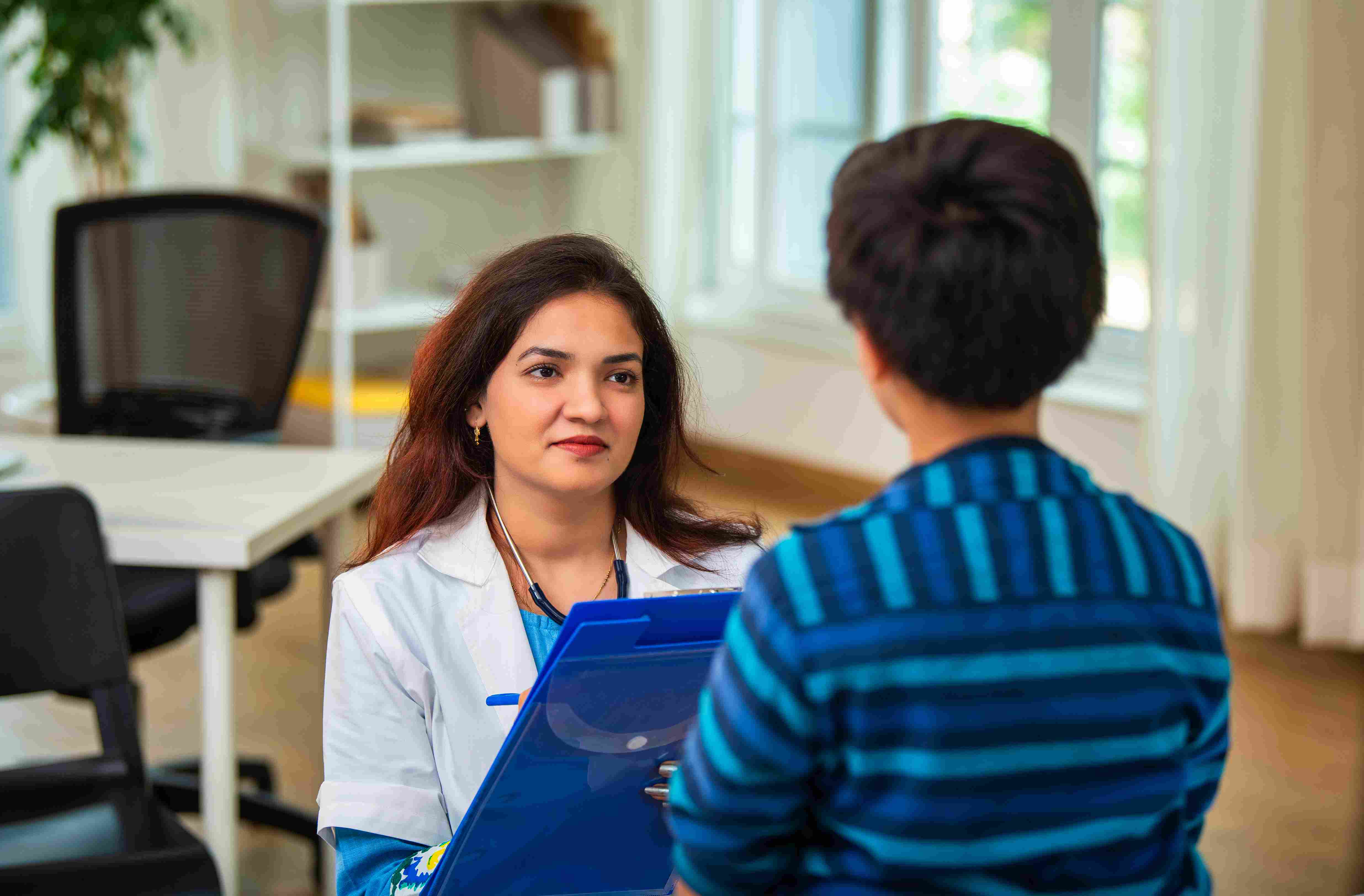 M&eacute;dica em consulta com crian&ccedil;a em ambiente cl&iacute;nico, com um sorriso atento e profissional, segurando prancheta.