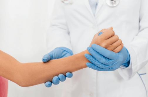 Profissional de sa&uacute;de com jaleco branco e luvas azuis segurando e examinando o punho de um(a) paciente.