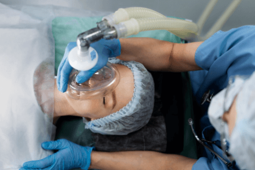 Profissional de sa&uacute;de prepara paciente para intuba&ccedil;&atilde;o, posicionando m&aacute;scara de ventila&ccedil;&atilde;o com oxig&ecirc;nio no rosto da paciente, que est&aacute; sedada e deitada em ambiente cir&uacute;rgico, usando touca hospitalar.