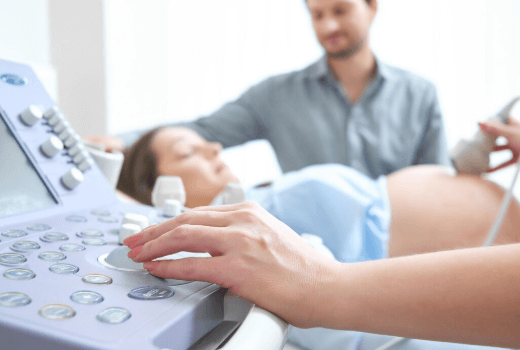 A imagem mostra uma mulher sendo submetida a um exame de ultrassonografia obst&eacute;trica, provavelmente para monitorar a gesta&ccedil;&atilde;o. A presen&ccedil;a do acompanhante ao fundo sugere que ele est&aacute; l&aacute; para apoiar a mulher durante o procedimento. O foco est&aacute; no aparelho de ultrassom, que &eacute; usado para visualizar o beb&ecirc; e verificar o desenvolvimento da gravidez.