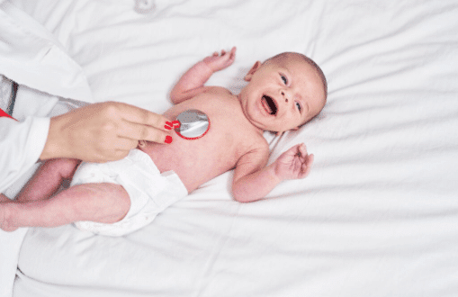 Um beb&ecirc; deitado de fralda sobre um len&ccedil;ol branco, sendo examinado por um profissional de sa&uacute;de com um estetosc&oacute;pio colocado no peito do beb&ecirc;.