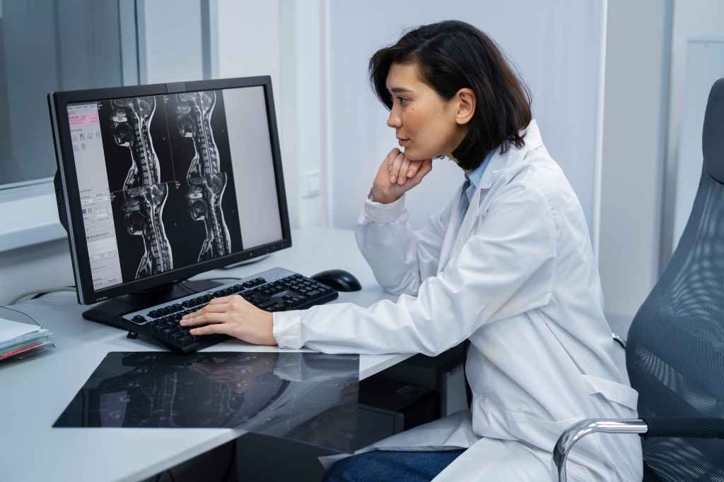 M&eacute;dica sentada em consult&oacute;rio analisando imagens de resson&acirc;ncia magn&eacute;tica da coluna vertebral exibidas em um monitor de computador.