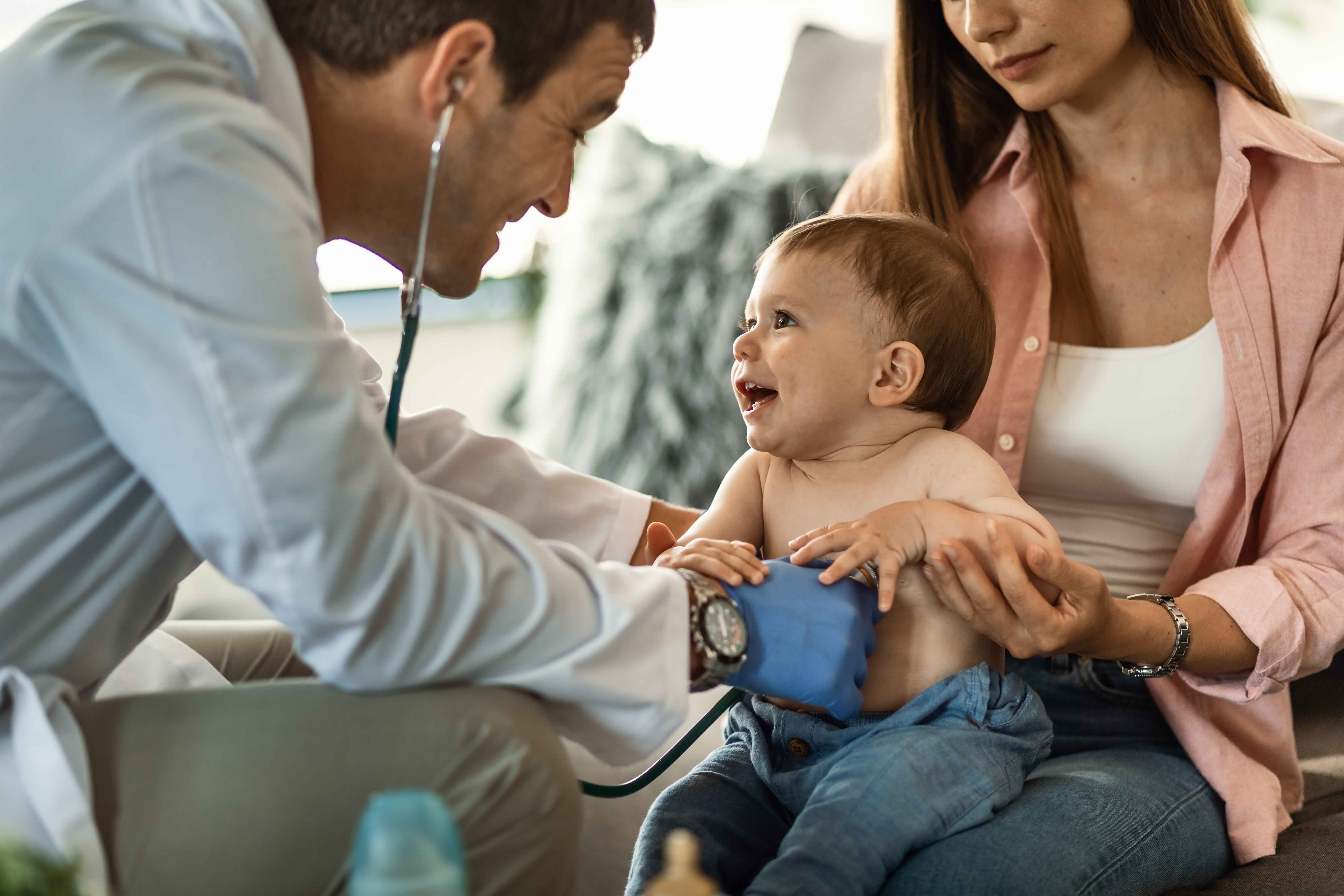 M&eacute;dico pediatra auscultando o cora&ccedil;&atilde;o de um beb&ecirc; sorridente no colo da m&atilde;e durante consulta.