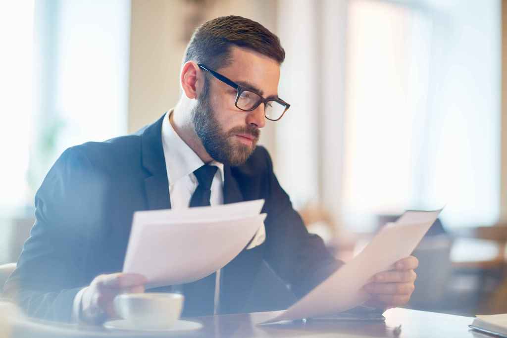 Homem de terno e &oacute;culos analisando documentos com aten&ccedil;&atilde;o, sentado &agrave; mesa com uma x&iacute;cara de caf&eacute; ao lado, em ambiente profissional iluminado.