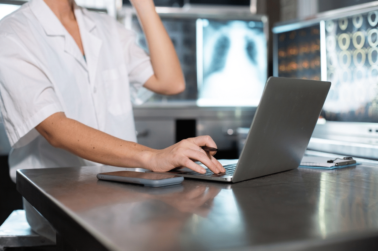 Radiologista utilizando notebook em sala de diagn&oacute;stico por imagem com exames exibidos em monitores ao fundo.