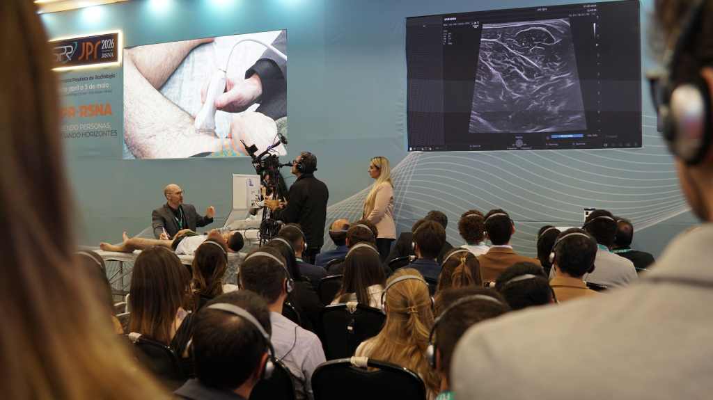 Dr. Everaldo Gregio Junior, vestindo terno cinza, gesticula durante uma aula pr&aacute;tica de ultrassonografia musculoesquel&eacute;tica na JPR 2026. Ele est&aacute; sentado pr&oacute;ximo a um paciente deitado, e um c&acirc;mera e uma assistente est&atilde;o ao seu lado. O p&uacute;blico assiste atentamente.