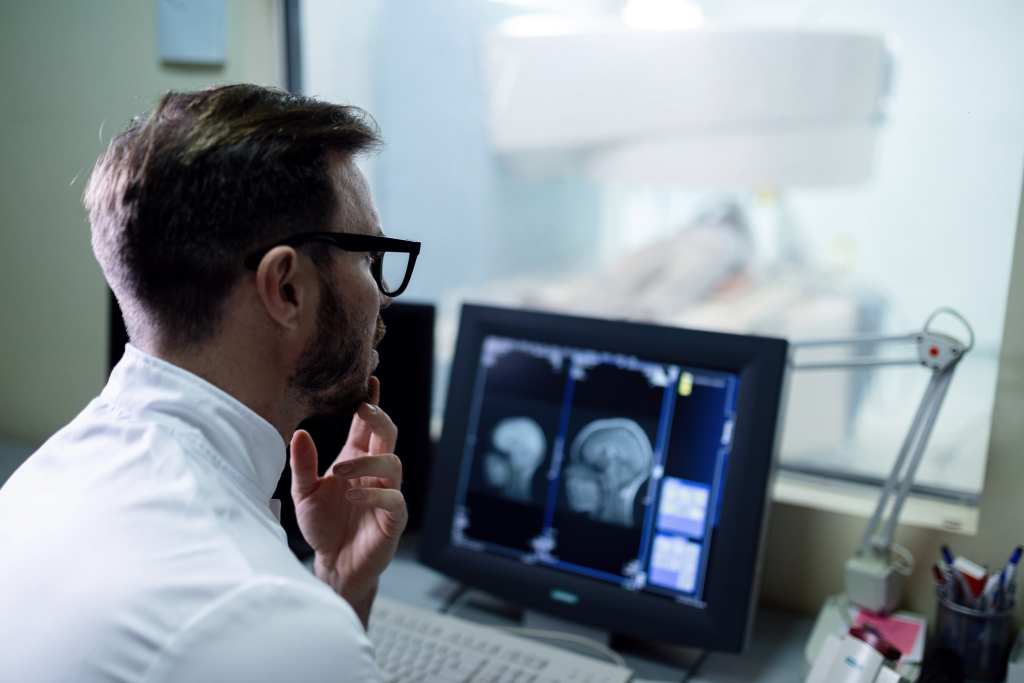 M&eacute;dico radiologista analisando exames de resson&acirc;ncia magn&eacute;tica em monitor dentro de ambiente hospitalar, com equipamento de diagn&oacute;stico por imagem ao fundo.