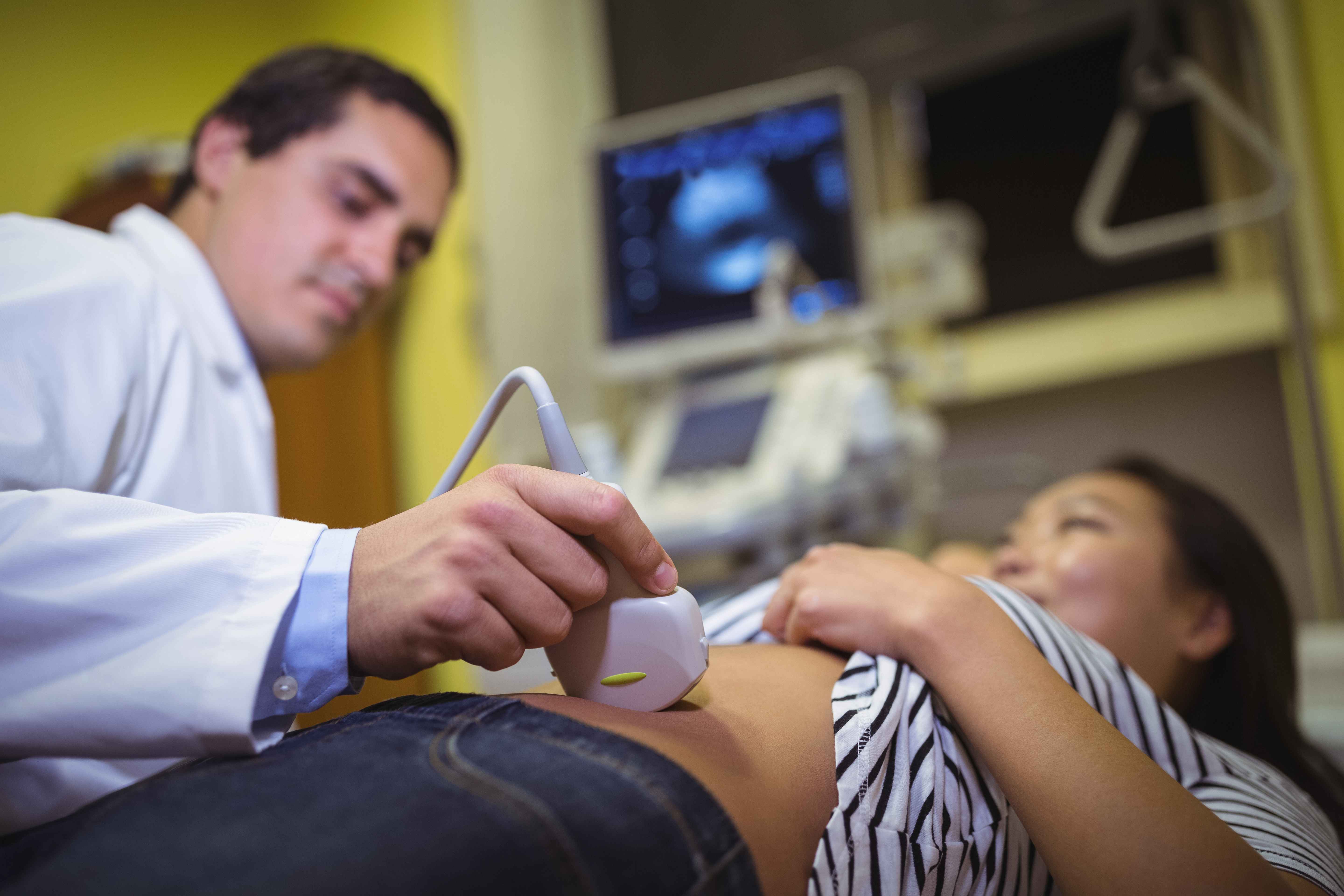 M&eacute;dico realiza exame de ultrassonografia em gestante deitada em maca, com aparelho de imagem ao fundo em ambiente cl&iacute;nico.