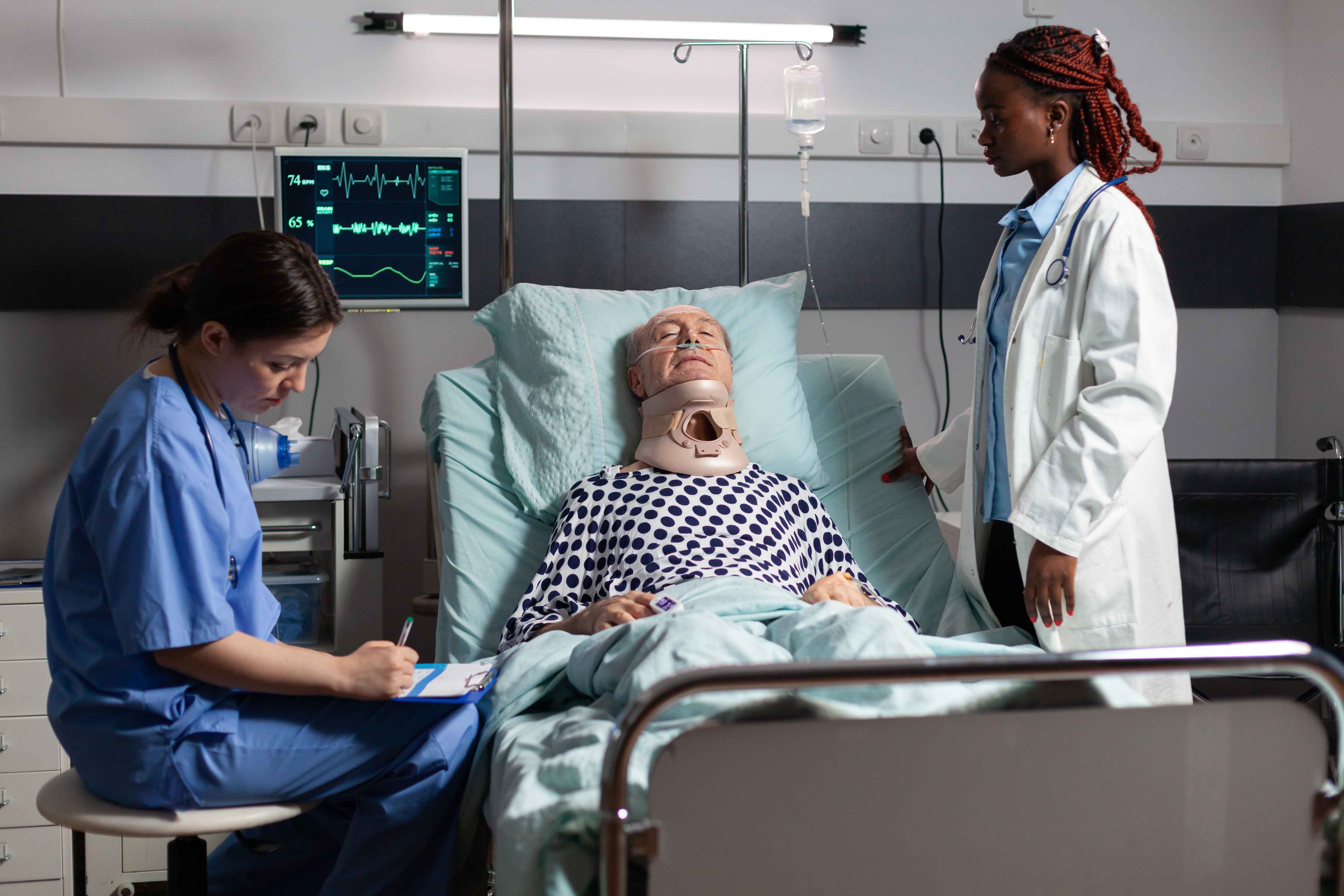 Paciente idoso internado em leito hospitalar, usando colar cervical e monitoriza&ccedil;&atilde;o card&iacute;aca, enquanto duas profissionais de sa&uacute;de realizam atendimento &agrave; beira do leito em ambiente de UTI.