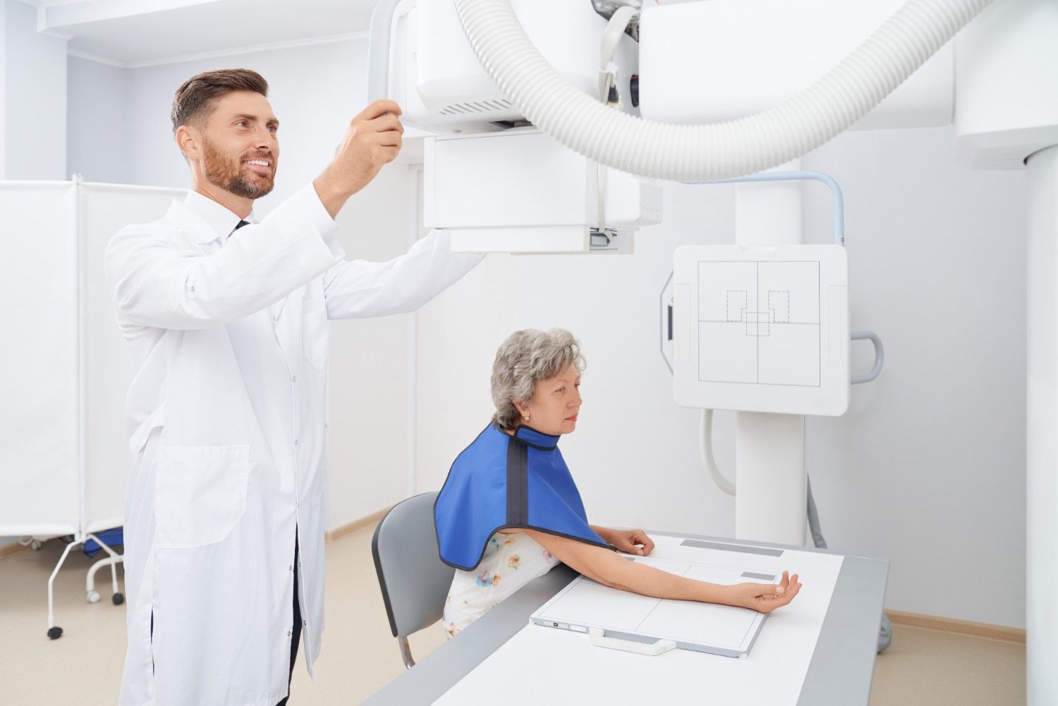 M&eacute;dico radiologista ajustando equipamento de radiografia enquanto paciente idosa realiza exame em cl&iacute;nica de diagn&oacute;stico por imagem.
