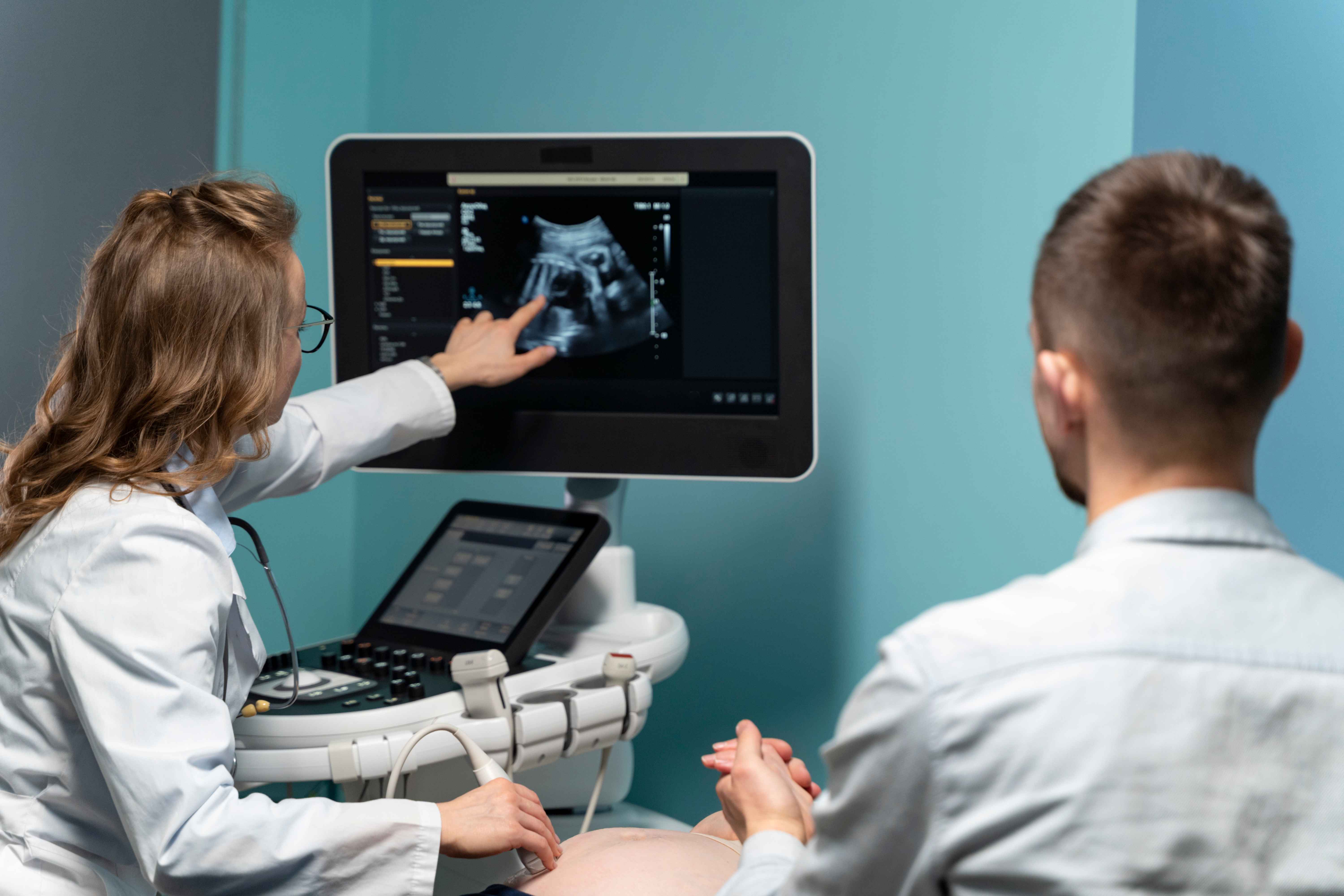 Uma profissional de sa&uacute;de usando jaleco branco aponta para uma imagem de ultrassom exibida em um monitor enquanto realiza ou explica um exame pr&eacute;-natal. Uma pessoa sentada, possivelmente um futuro pai ou acompanhante, observa a tela. O equipamento de ultrassom e o monitor est&atilde;o vis&iacute;veis em uma sala de exames com paredes em tons de azul claro.
