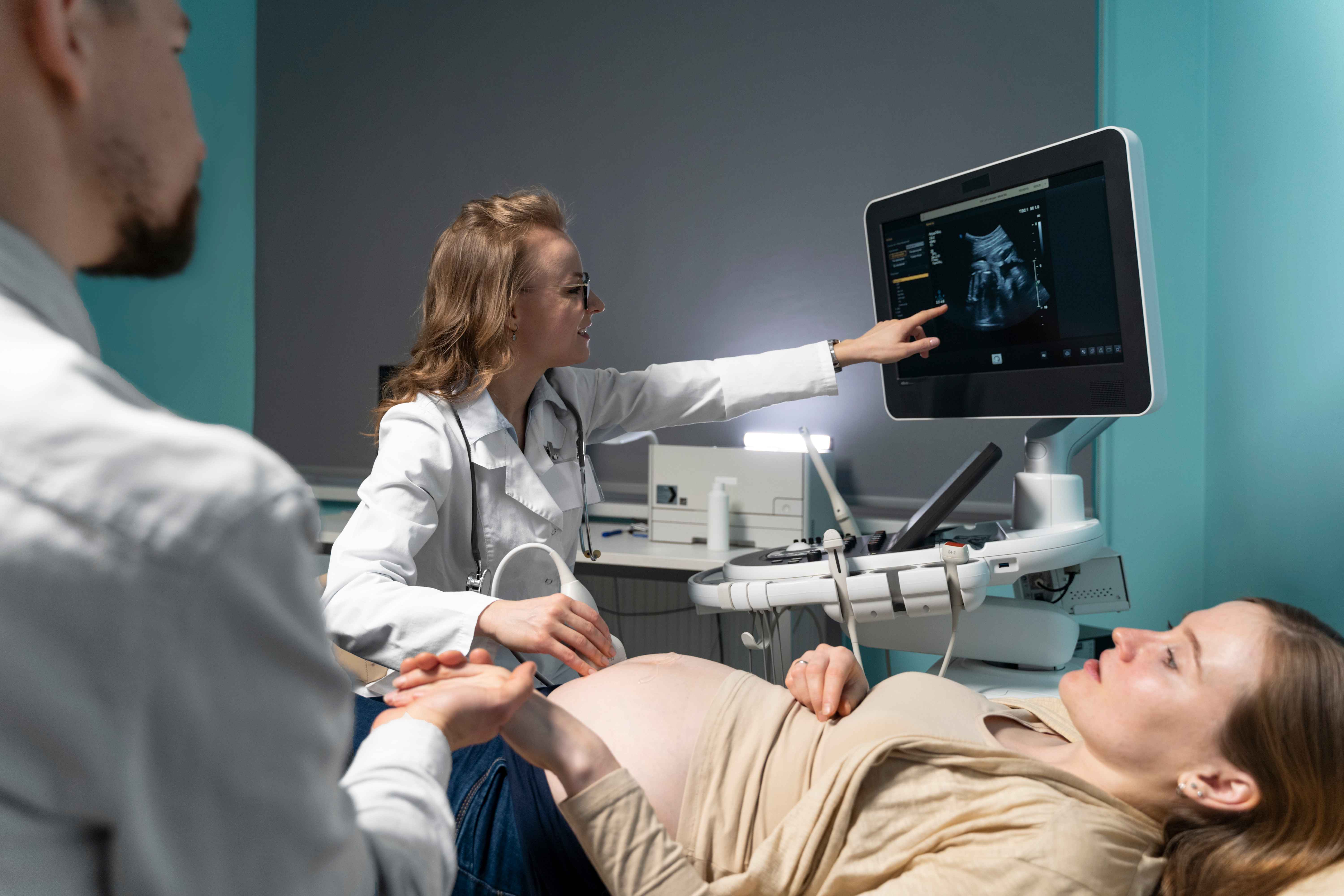 A imagem mostra uma consulta pr&eacute;-natal com exame de ultrassom. Uma m&eacute;dica est&aacute; ao lado de uma gestante deitada na maca, apontando para a tela do equipamento de ultrassom, onde &eacute; poss&iacute;vel ver a imagem do beb&ecirc;.