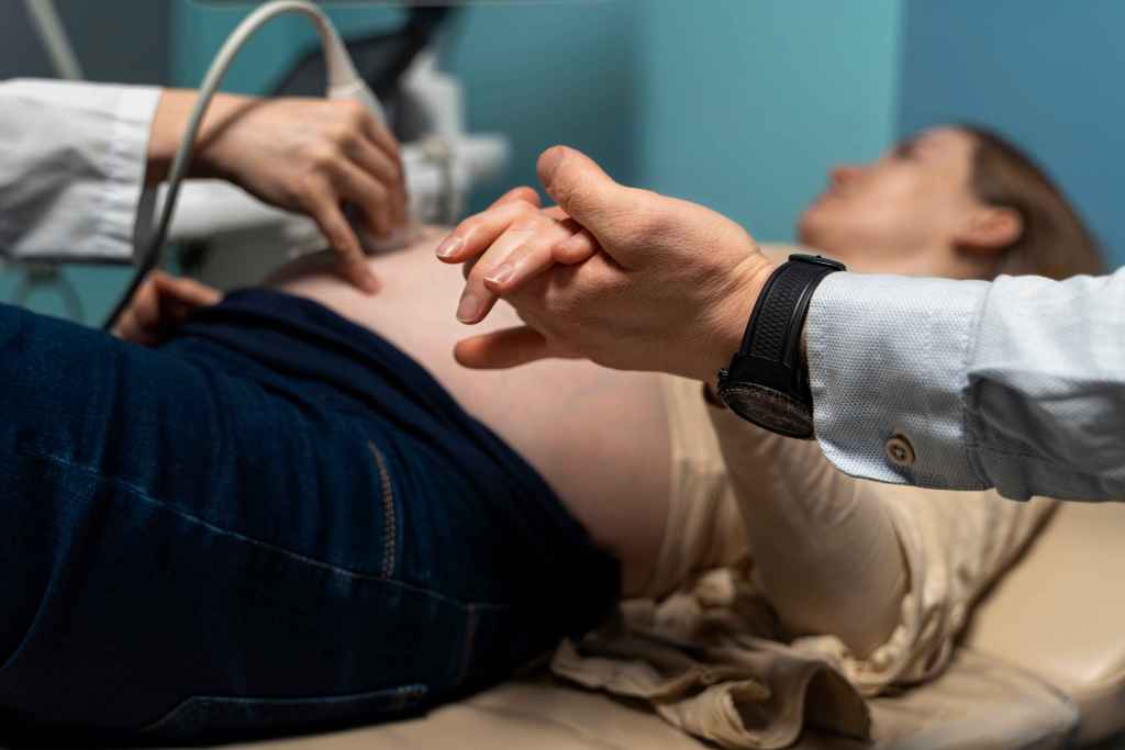 Profissional de sa&uacute;de realiza exame de ultrassonografia obst&eacute;trica em gestante deitada na maca, enquanto ela segura a m&atilde;o de um acompanhante durante o atendimento.