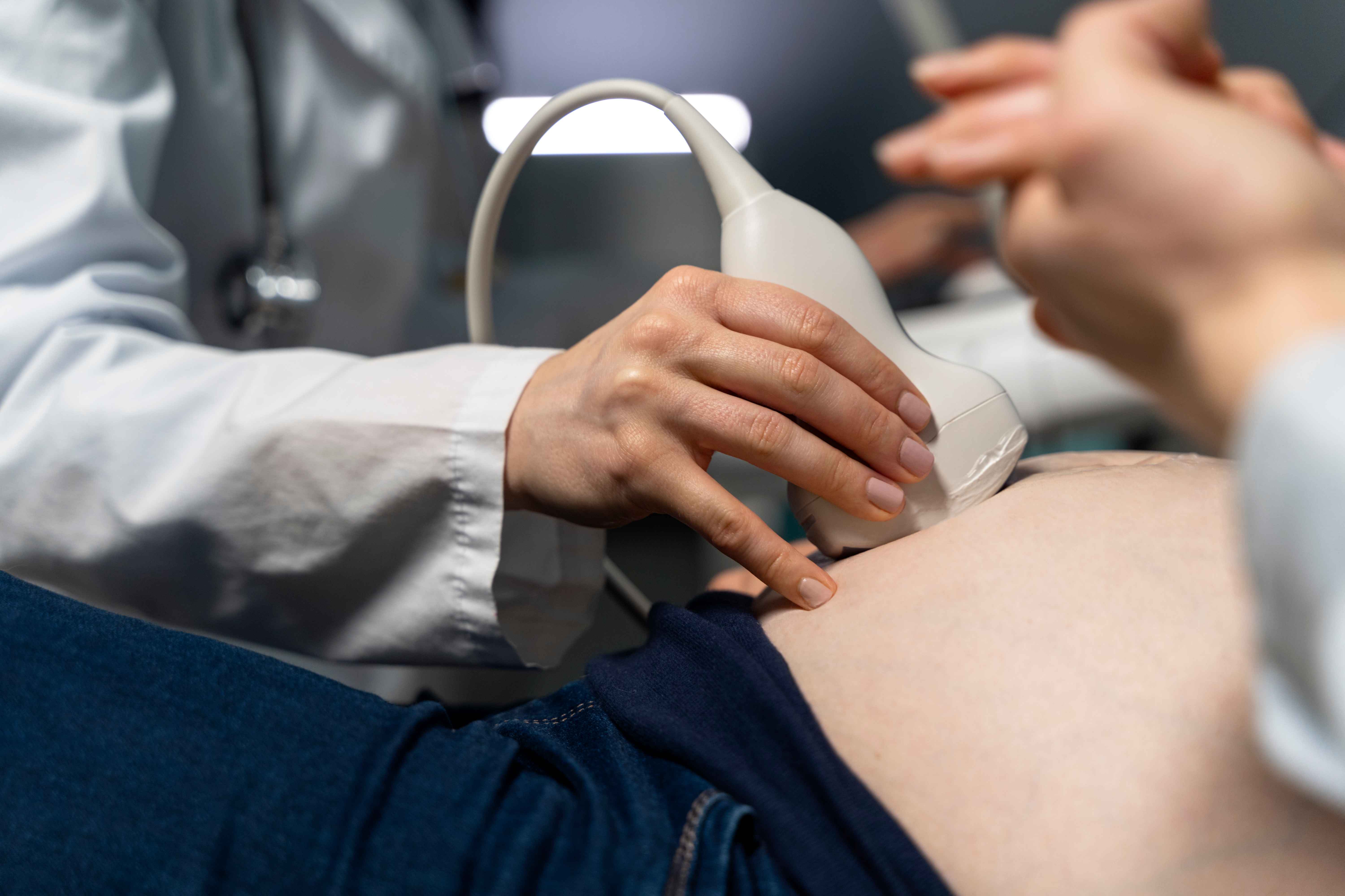 Profissional de sa&uacute;de realiza exame de ultrassonografia obst&eacute;trica em uma gestante, posicionando o transdutor sobre o abd&ocirc;men da paciente durante consulta de acompanhamento pr&eacute;-natal em ambiente cl&iacute;nico.