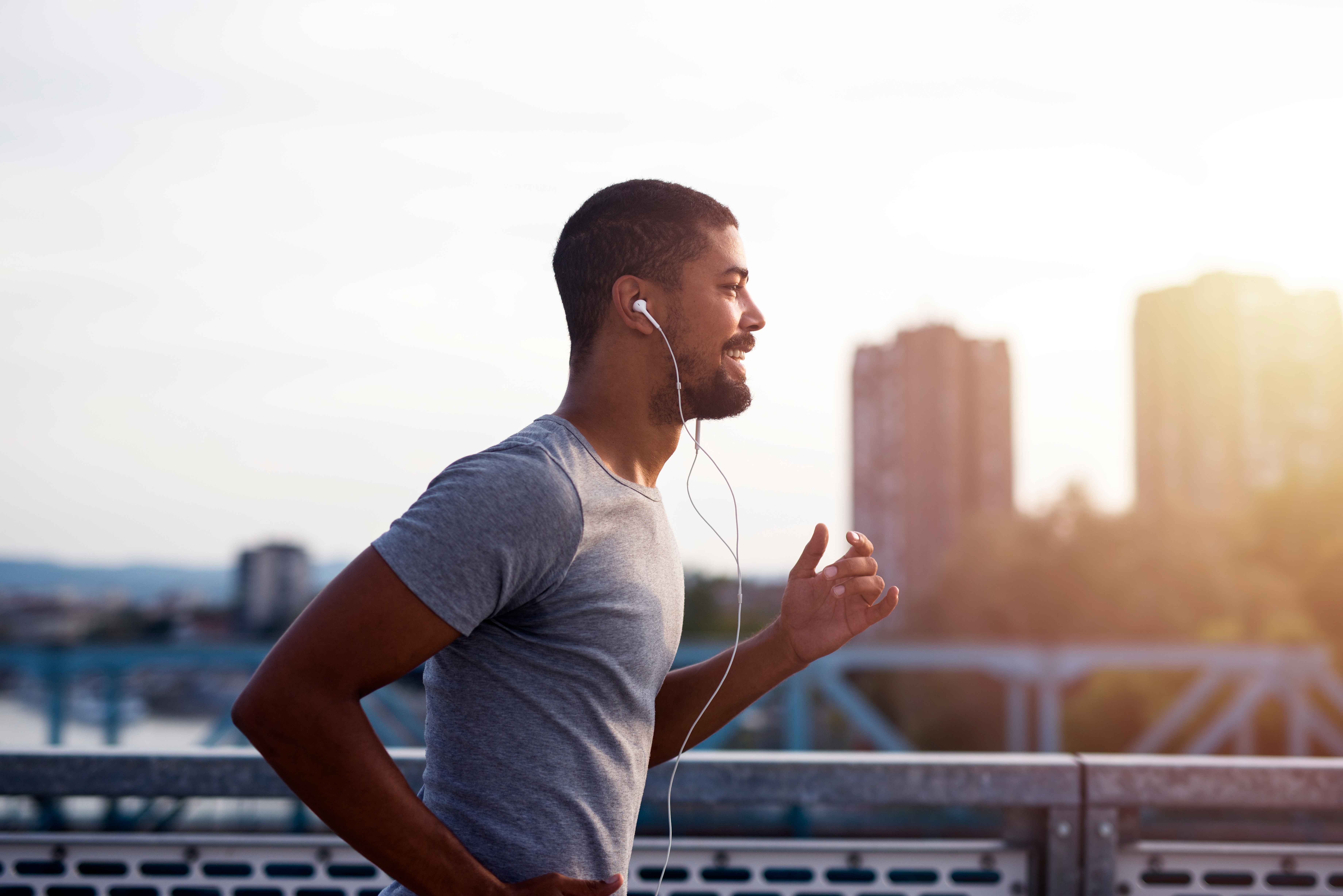 Homem correndo ao ar livre em uma ponte urbana durante o nascer ou p&ocirc;r do sol. Ele usa camiseta cinza e fones de ouvido com fio, enquanto pratica atividade f&iacute;sica com pr&eacute;dios desfocados ao fundo e luz natural suave iluminando a cena.