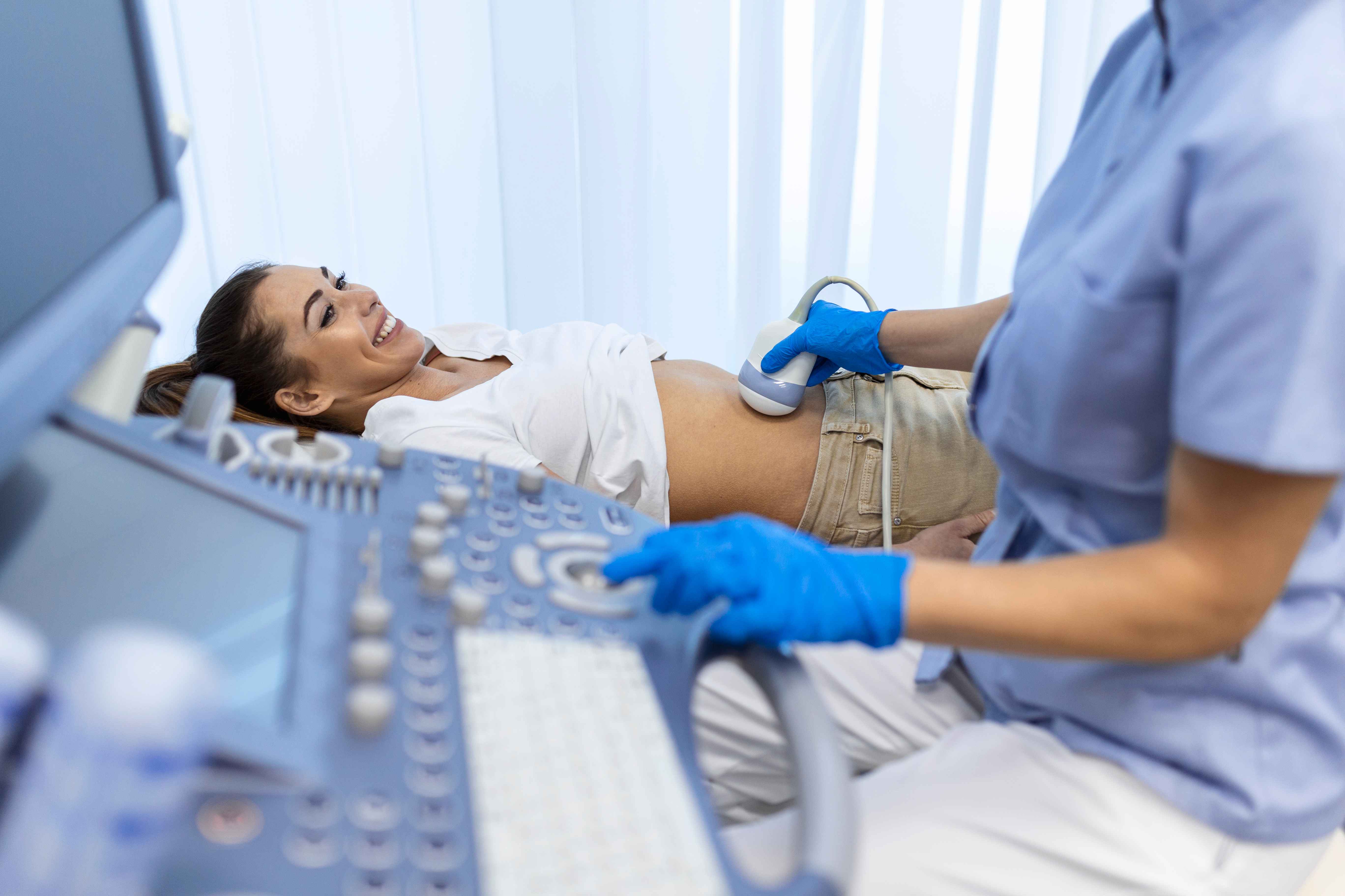 Mulher gr&aacute;vida sorrindo durante exame de ultrassonografia obst&eacute;trica em ambiente cl&iacute;nico. Um profissional de sa&uacute;de utiliza o transdutor sobre o abd&ocirc;men da paciente enquanto opera o equipamento de ultrassom, cuja tela aparece parcialmente ao lado.
