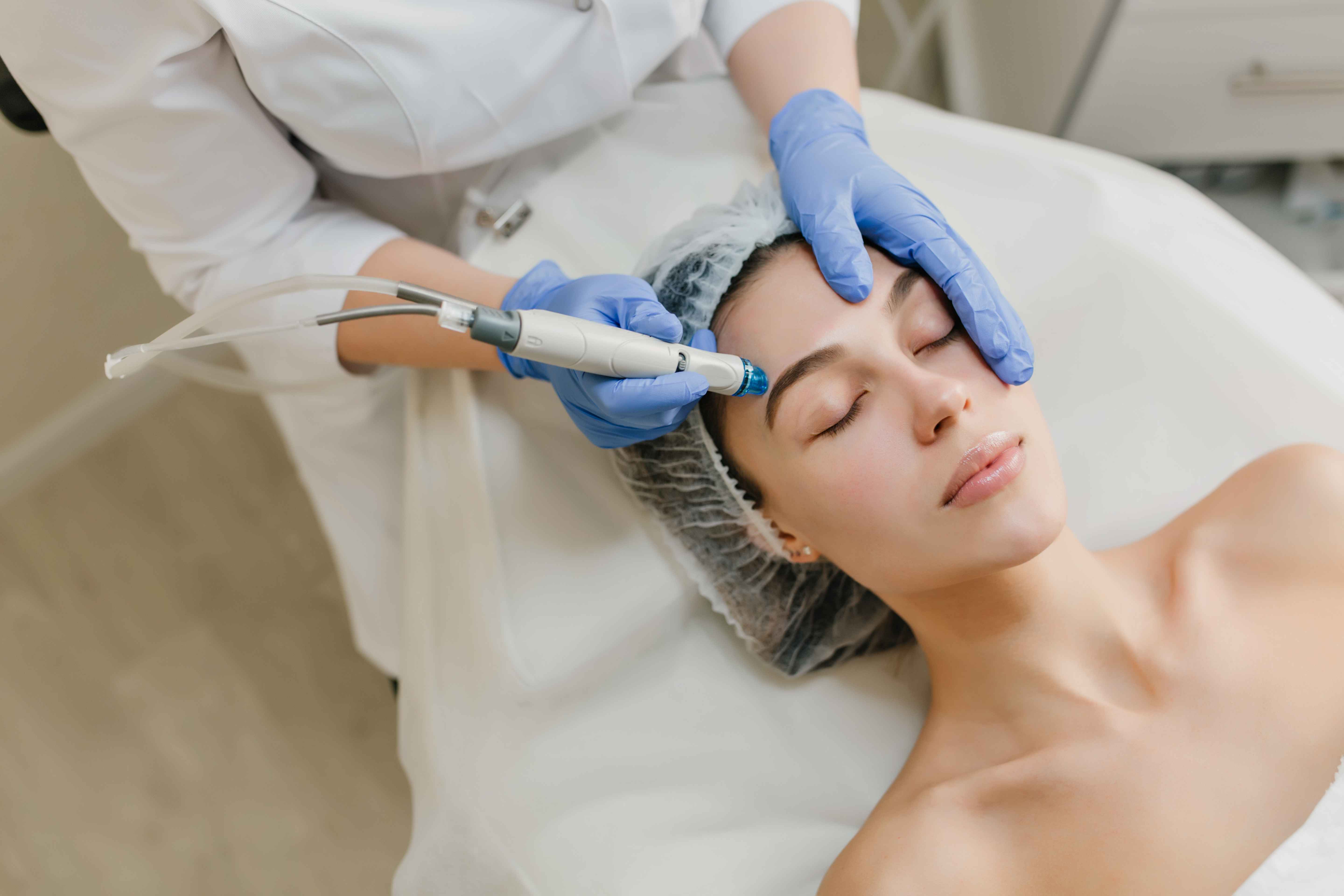 Mulher deitada em maca durante procedimento est&eacute;tico facial realizado por profissional de sa&uacute;de com luvas azuis. Um equipamento &eacute; aplicado na regi&atilde;o da testa e sobrancelhas, em ambiente cl&iacute;nico, sugerindo tratamento dermatol&oacute;gico ou de rejuvenescimento da pele.