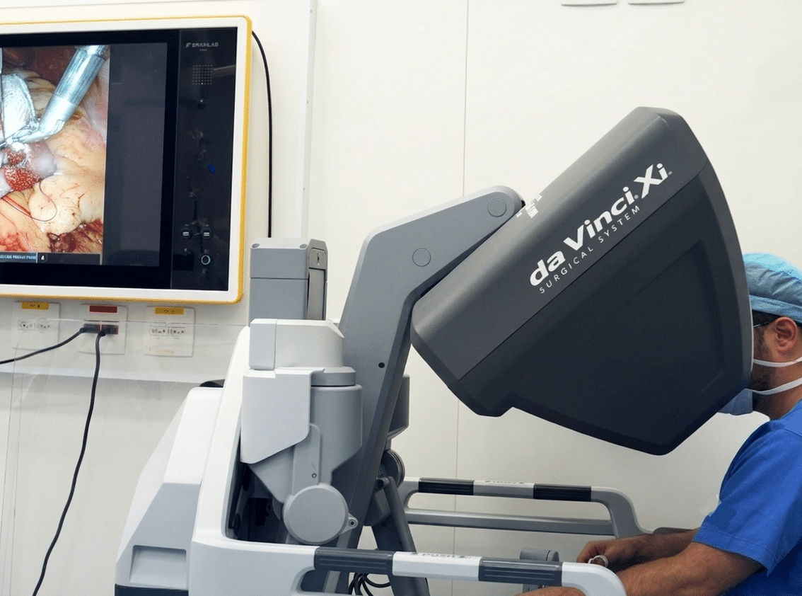 Imagem de um sistema rob&oacute;tico cir&uacute;rgico da Vinci Xi em sala de cirurgia, com m&eacute;dico operando e monitor exibindo o campo cir&uacute;rgico.
