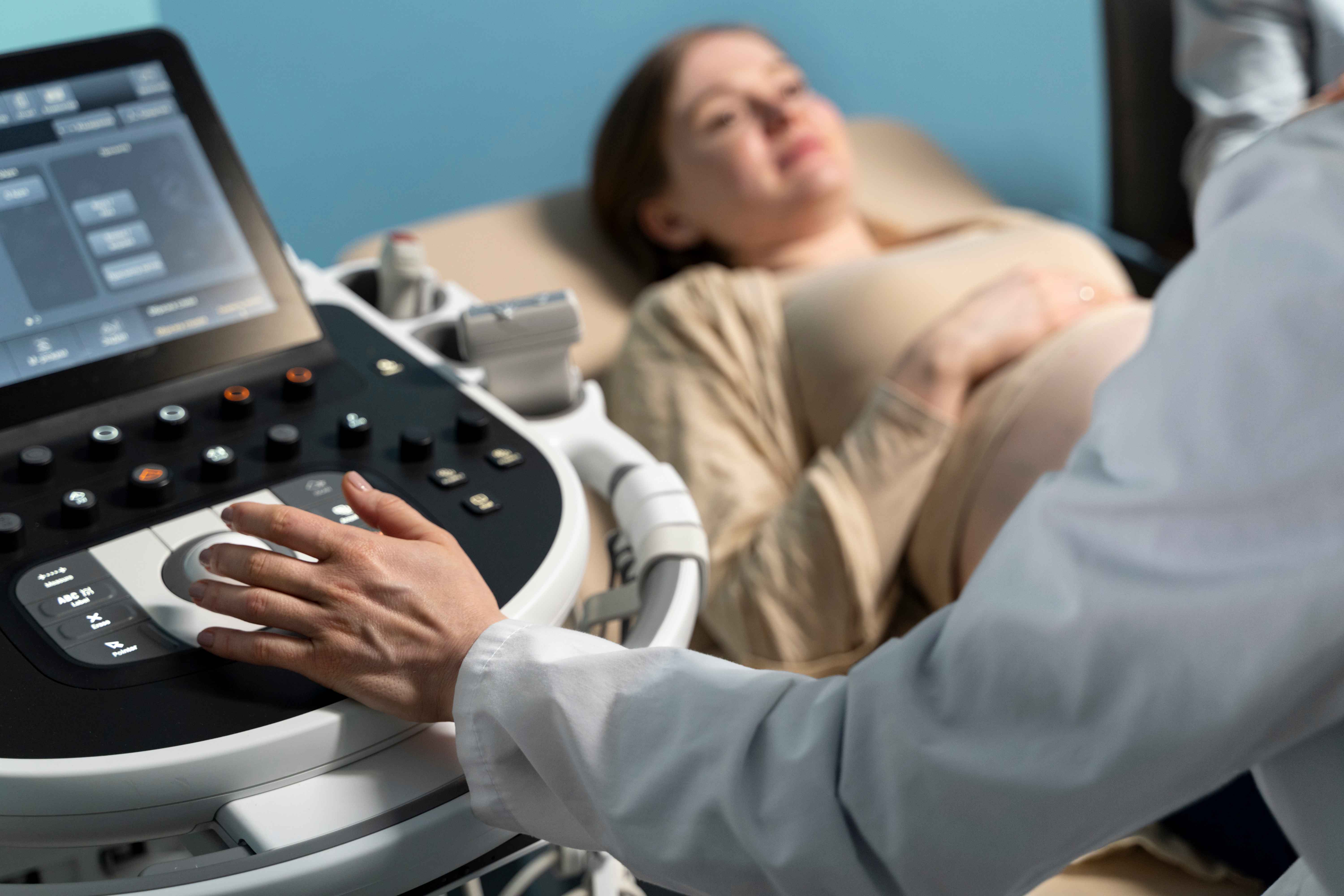 M&eacute;dico realiza exame de ultrassonografia obst&eacute;trica em gestante deitada em maca. Em primeiro plano, aparece o painel do equipamento de ultrassom e a m&atilde;o do profissional ajustando os controles. Ao fundo, a paciente gr&aacute;vida repousa confortavelmente durante a avalia&ccedil;&atilde;o pr&eacute;-natal em ambiente cl&iacute;nico.