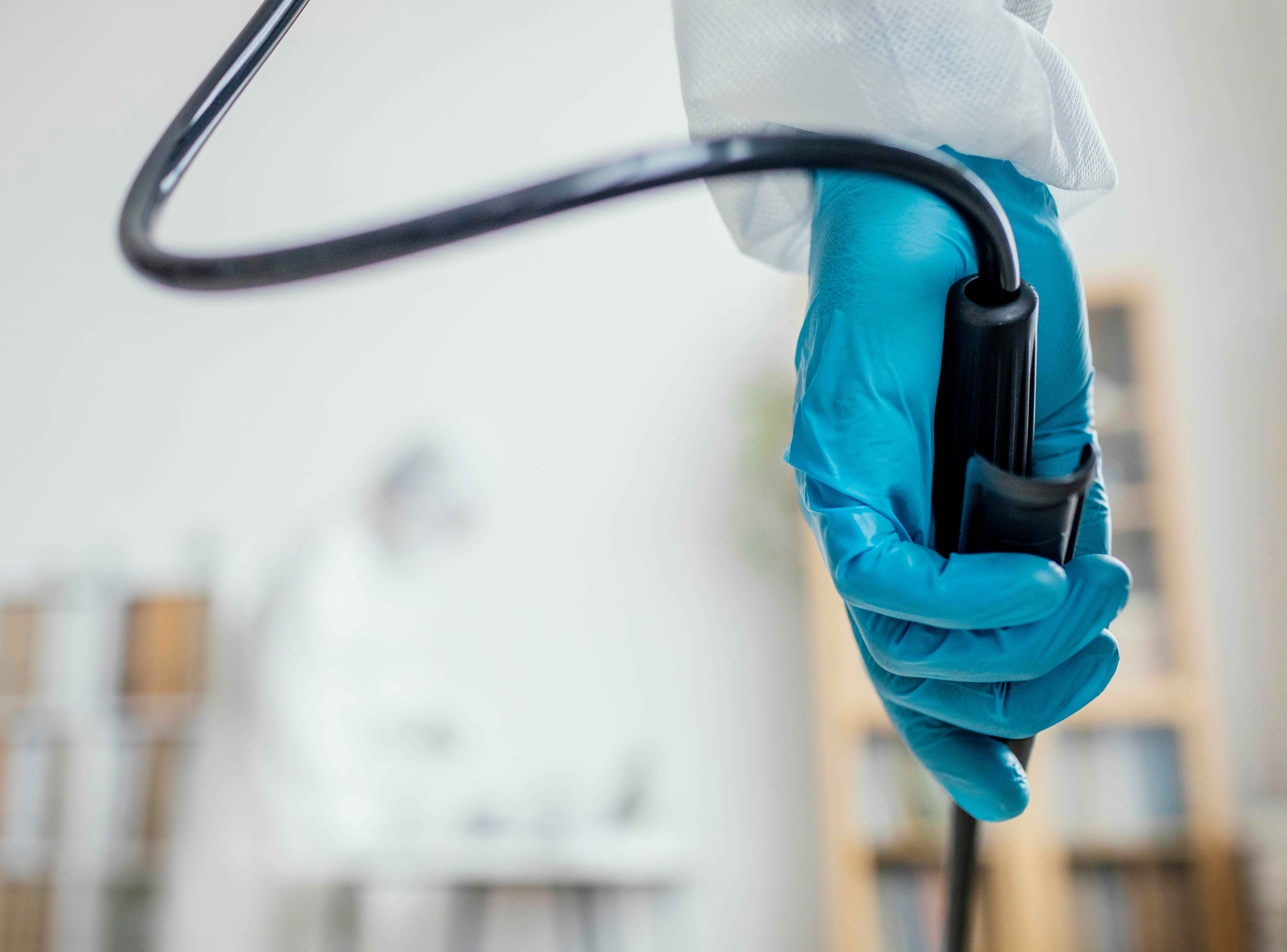 Profissional de sa&uacute;de utilizando luva azul segura um colonosc&oacute;pio em primeiro plano, com foco no cabo do equipamento. Ao fundo, desfocado, observa-se uma sala de endoscopia preparada para procedimentos diagn&oacute;sticos e terap&ecirc;uticos do trato gastrointestinal.