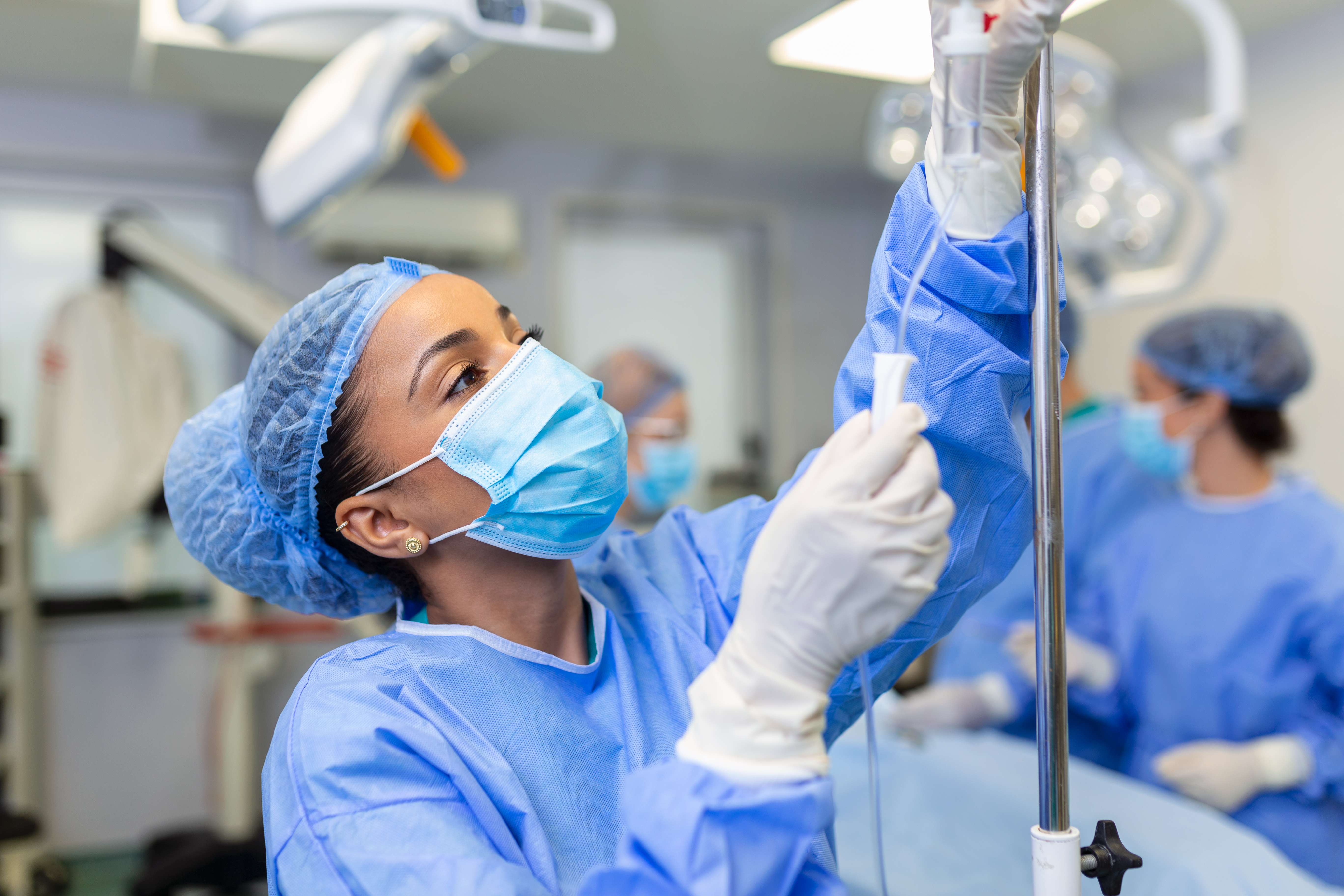 Profissional de sa&uacute;de em centro cir&uacute;rgico ajustando uma medica&ccedil;&atilde;o em acesso intravenoso (soro) durante procedimento cir&uacute;rgico, usando m&aacute;scara, touca e luvas, com equipe m&eacute;dica desfocada ao fundo.