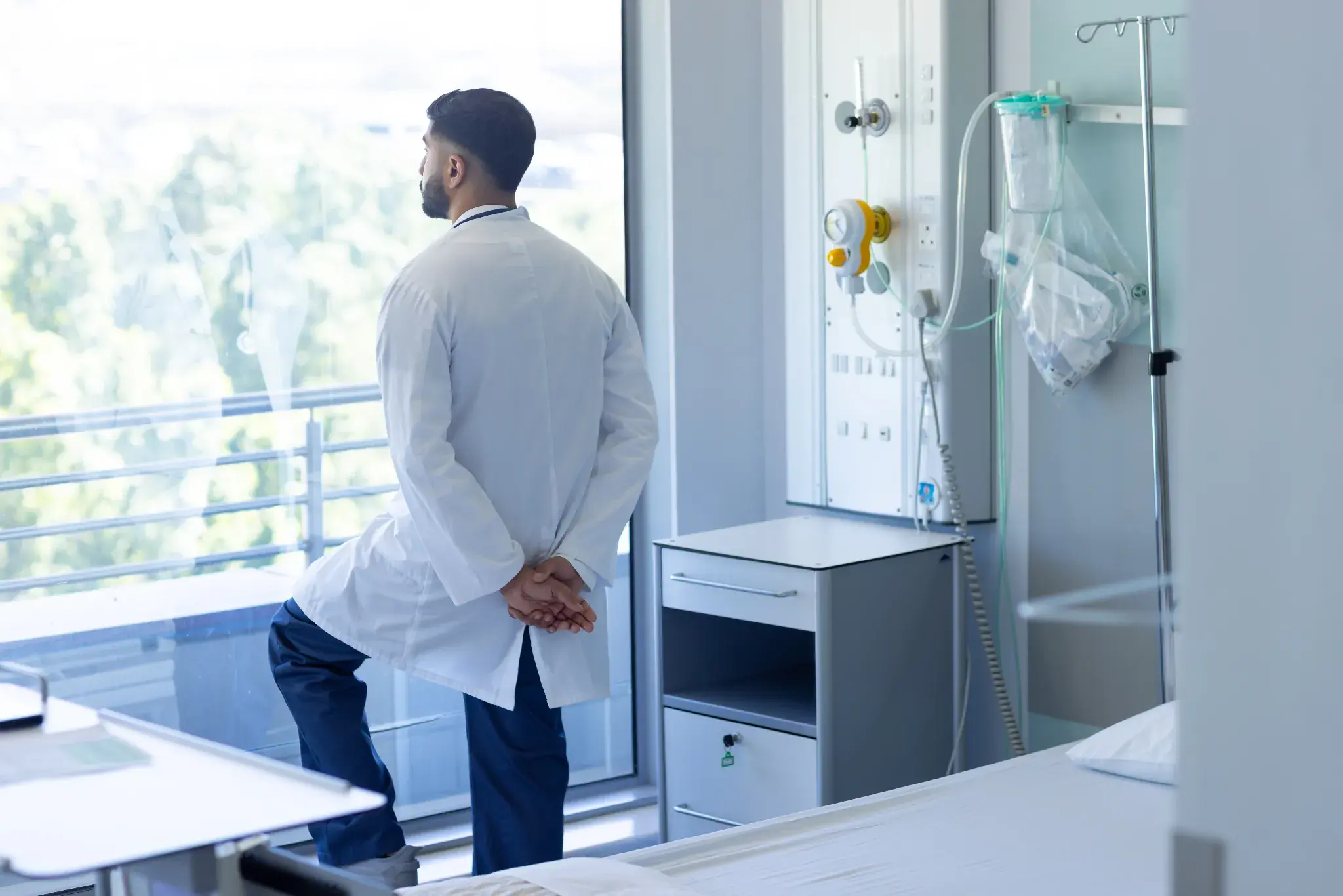 M&eacute;dico de costas, usando jaleco branco, observa pela janela de um quarto hospitalar.