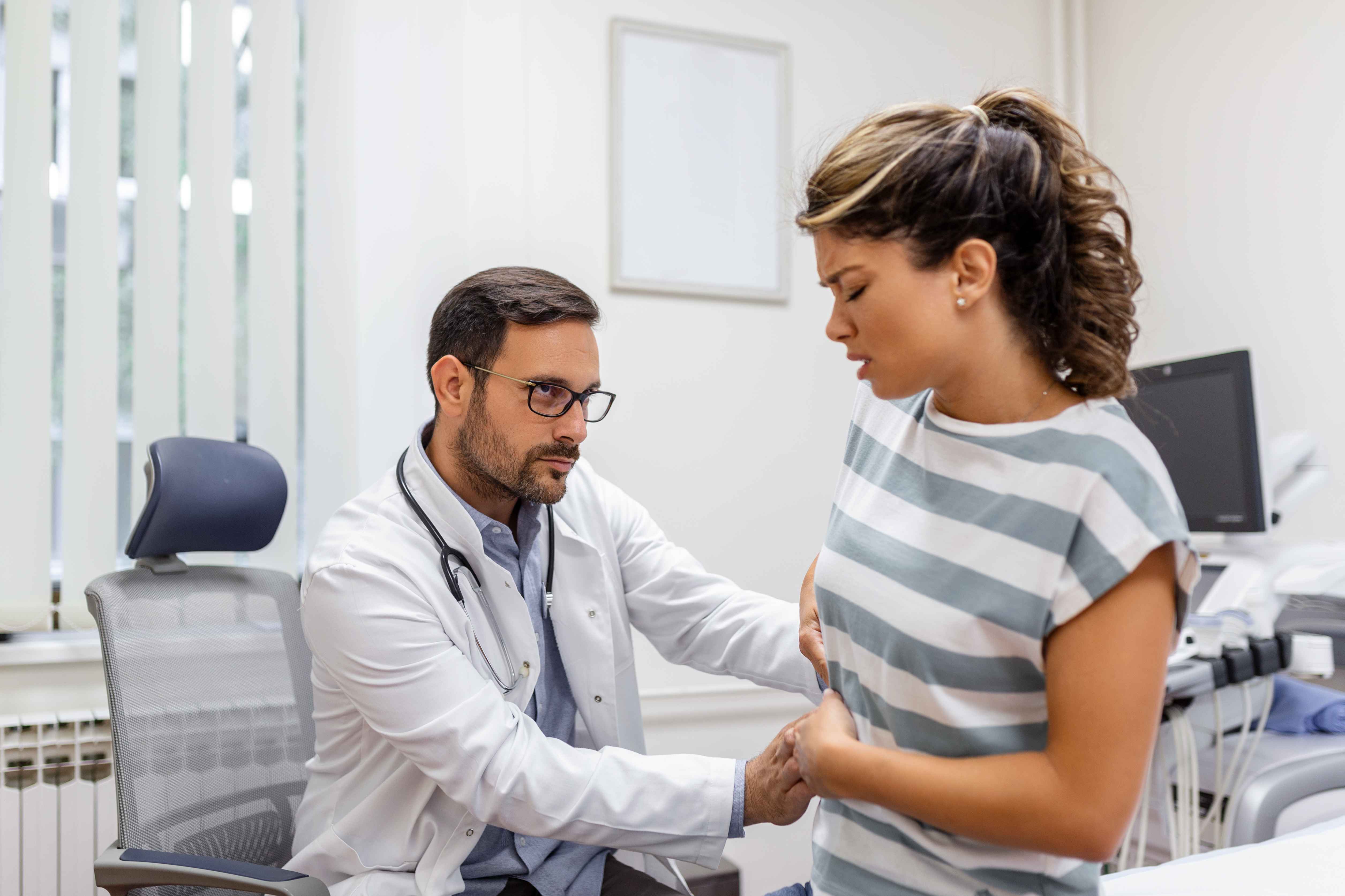 Mulher jovem em consulta m&eacute;dica segura o lado direito do abd&ocirc;men enquanto um m&eacute;dico sentado realiza exame f&iacute;sico na regi&atilde;o abdominal, em um consult&oacute;rio iluminado com equipamentos m&eacute;dicos ao fundo.