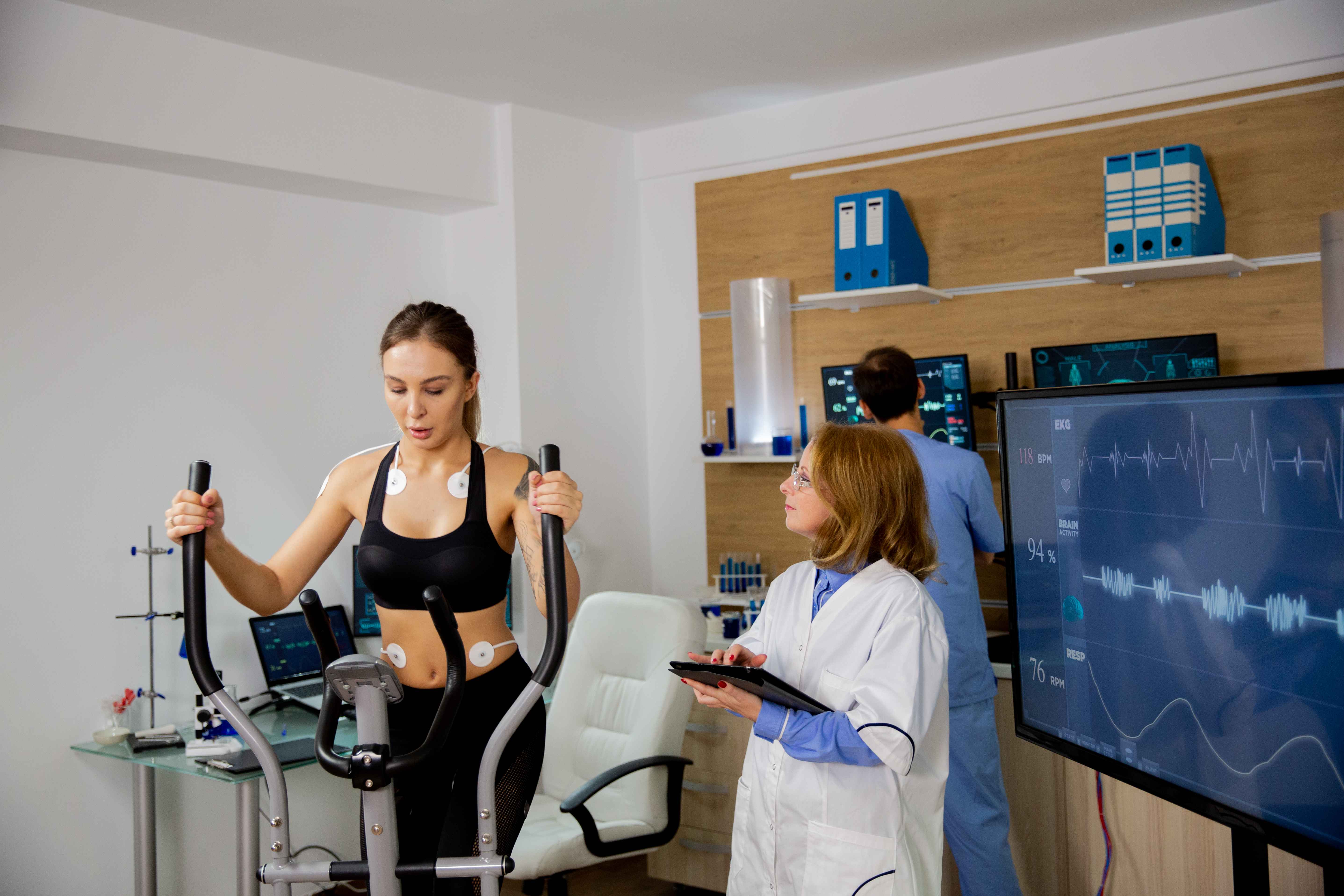 Atleta feminina realizando esforço físico no stepper enquanto o médico a acompanha. Laboratório de esportes.