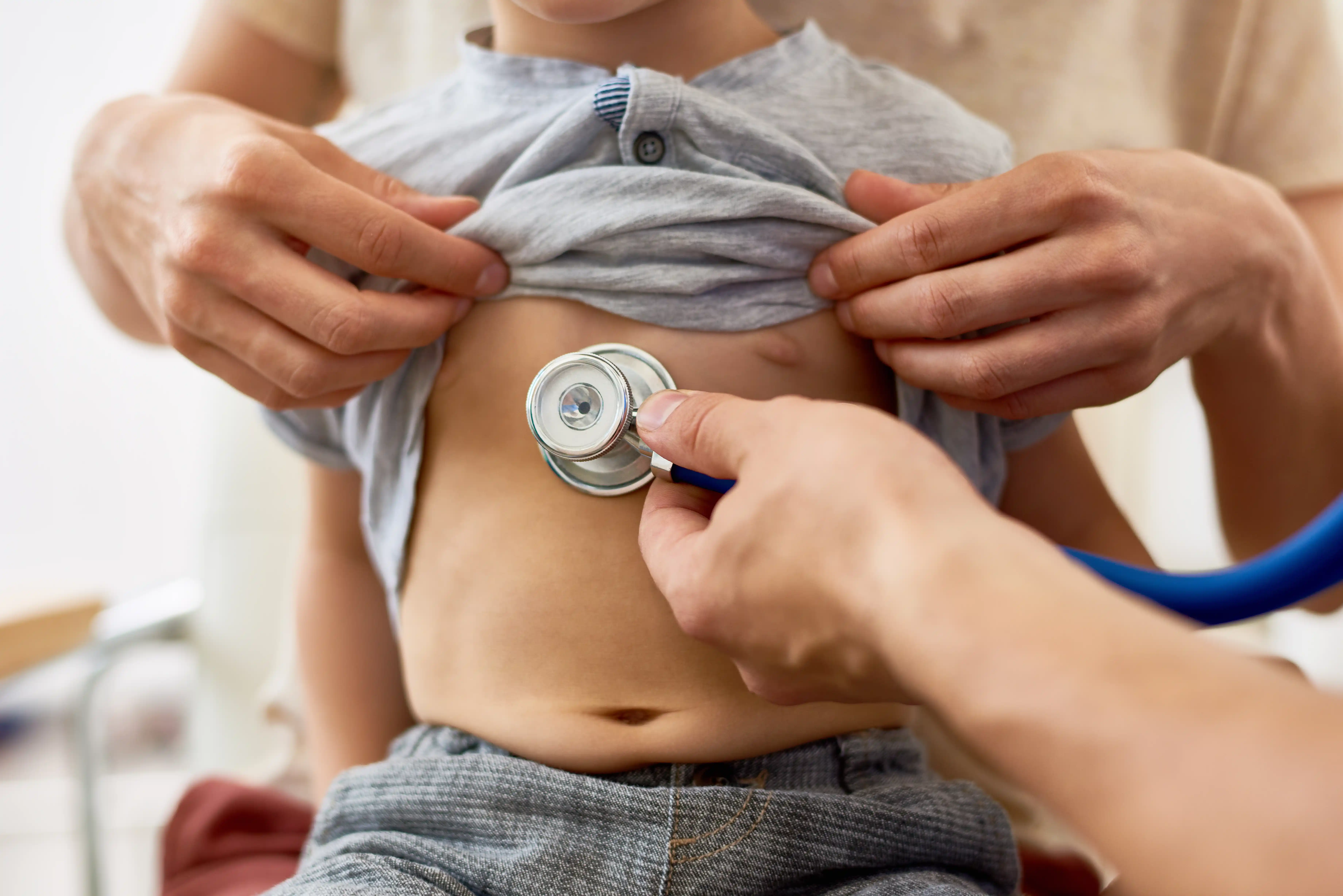 Médico auscultando o coração de uma criança com estetoscópio enquanto um adulto levanta a camiseta dela